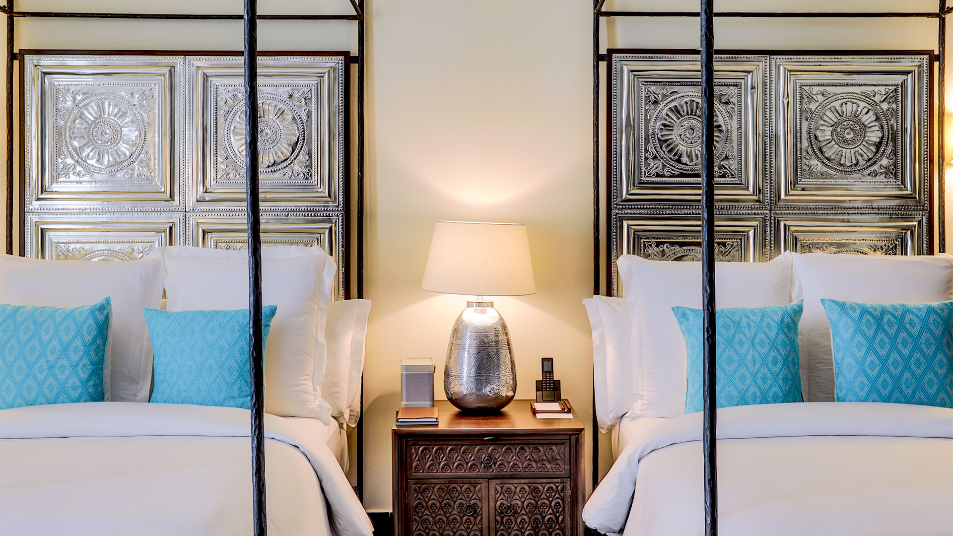 Urban junior suite double beds with ornate metal headboards flank a lamp on a table at Banyan Tree Puebla.