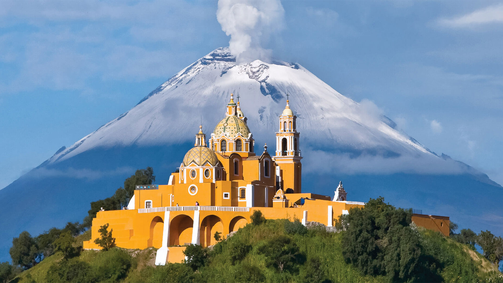 Church standing atop a hill, beneath a steaming volcano, showcasing Banyan Tree Puebla's scenic allure you can visit.