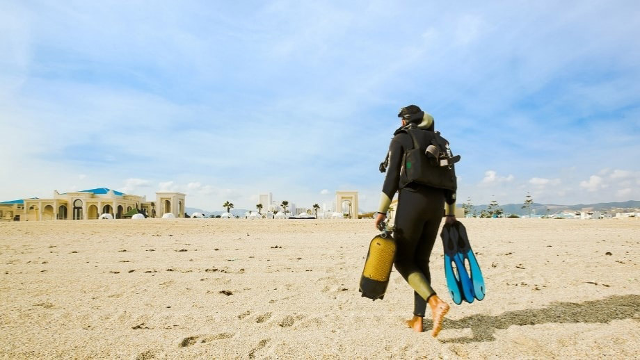 Diver walking on Banyan Tree Tamouda Bay beach, carrying gear for watersports activity.